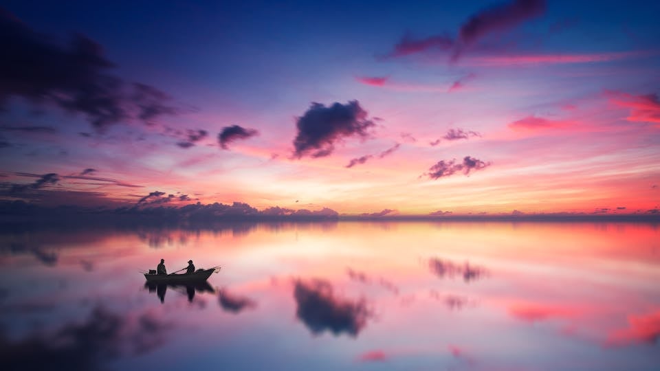 A tranquil sunset scene with a boat silhouetted against a vibrant sky reflected on a calm lake.